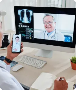Doctor using smartphone and desktop for video consultation with another physician—telemedicine platform displaying X-ray scans and AI-powered remote diagnostics.